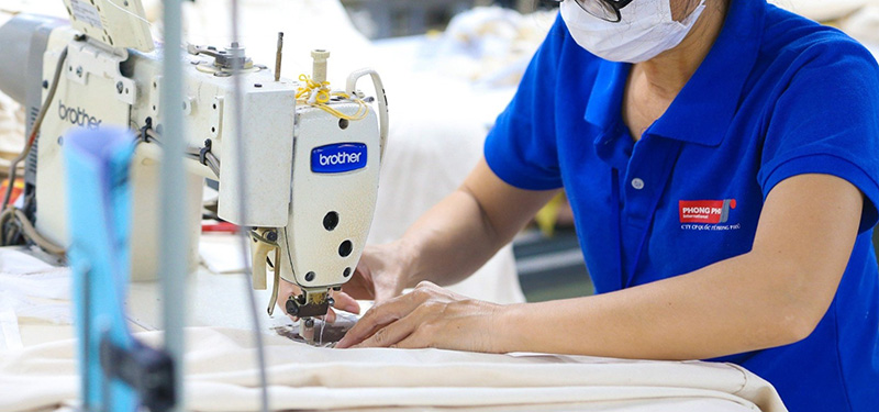 Worker in blue polo shirt operating industrial sewing machine in garment factory.