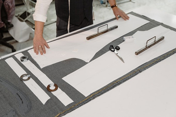 Tailor using pattern pieces, rulers, and scissors on gray fabric to cut clothing sections accurately.
