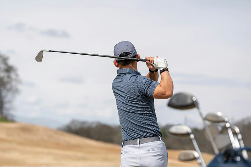 Golfer in striped blue polo shirt finishing swing on course beside golf bag.