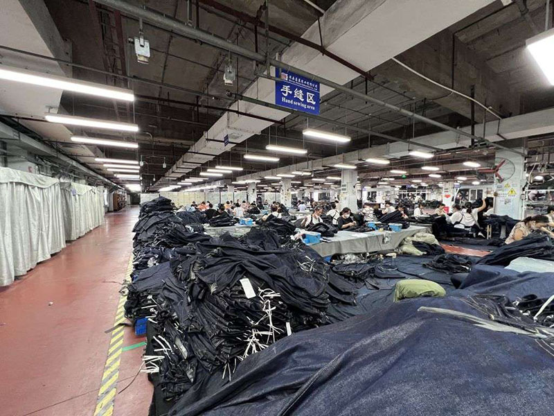 Large factory floor with rows of workers seated at tables, hand-sewing garments. Piles of dark fabric are stacked in the foreground under a sign reading "Hand sewing area."