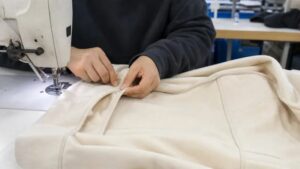 Garment worker sewing cream hoodie fabric on industrial machine during apparel manufacturing process