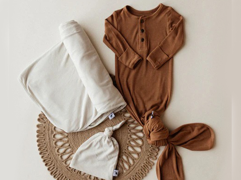 Baby outfit set including a brown long-sleeve knotted gown, a matching knotted hat, and a soft cream-colored blanket, all arranged on a round woven mat.