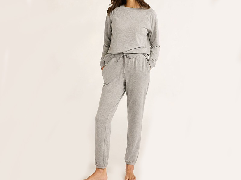 Woman wearing a light gray loungewear set with long sleeves and matching jogger-style pants with pockets, standing barefoot.