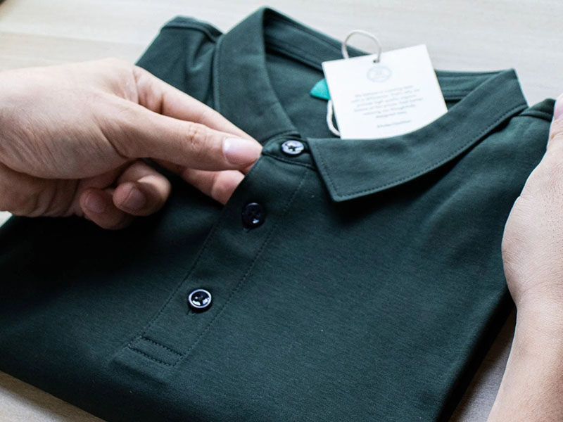 Hands holding a neatly folded dark green polo shirt with a tag attached, focusing on the buttons and collar.