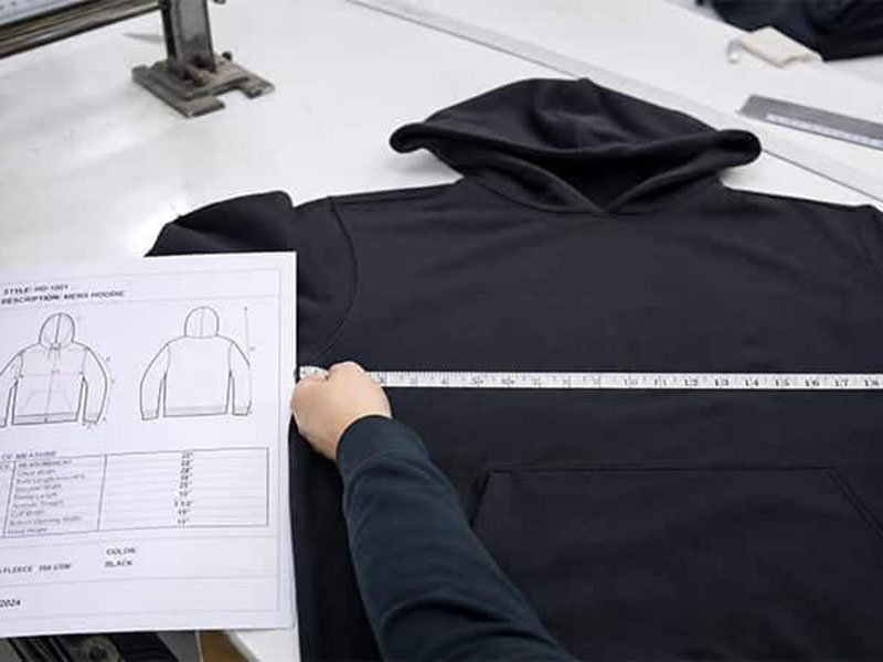Worker measuring width of a black hoodie with tape while referencing technical drawing for size verification
