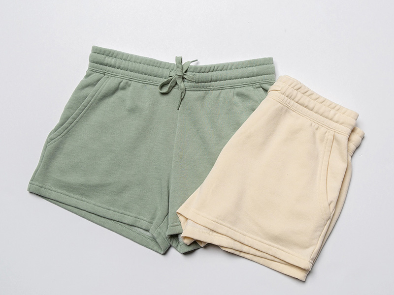 Two pairs of casual shorts laid flat on a white background. One pair is light green with a drawstring waist and side pockets, and the other pair is cream-colored with an elastic waistband and side pockets, folded neatly.