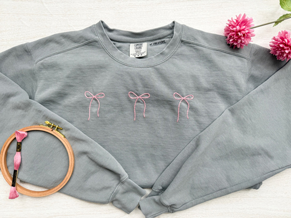 Gray sweatshirt with pink bow embroidery laid flat beside flowers and embroidery hoop.
