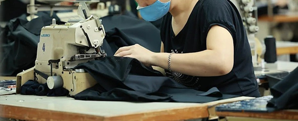 Garment worker sewing black fabric on an industrial machine while wearing a face mask.