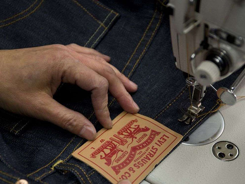Close-up of a person’s hand guiding denim fabric under a sewing machine, attaching the leather Levi Strauss & Co. patch on the back of a pair of jeans.