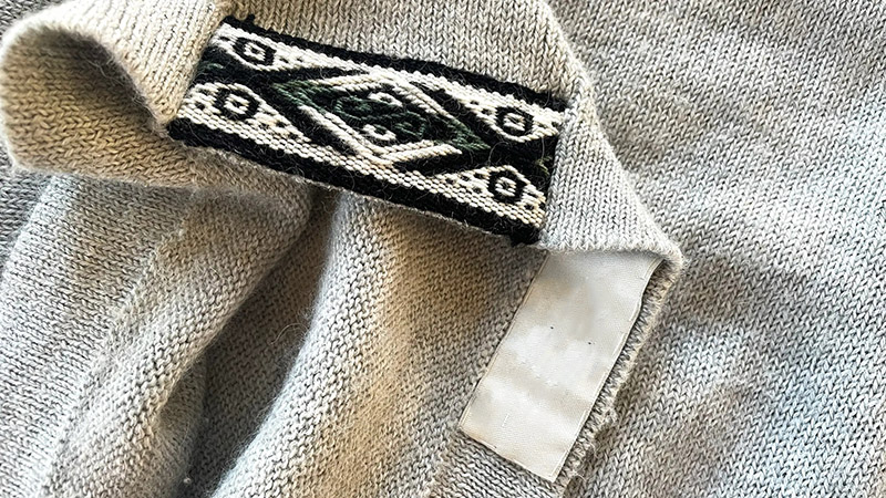 Close-up of a gray knitted garment with a woven black and white geometric label and a blank white care tag underneath.