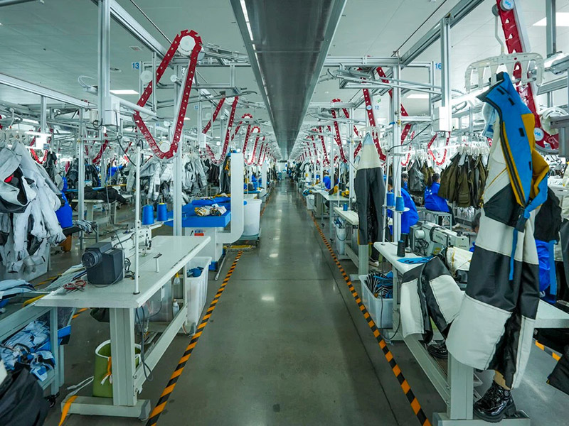 Long view down the center aisle of a large garment factory. Sewing stations line both sides of the aisle, each equipped with sewing machines, partially assembled garments, and fabric rolls.