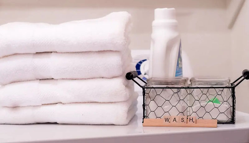 Stacked white towels beside laundry detergent and wash supplies on a clean surface.