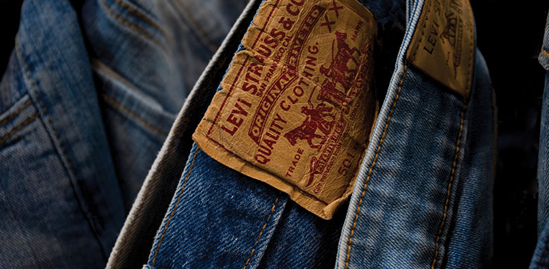 Close-up of a Levi's jeans leather patch on blue denim, showing the brand name and classic two-horse logo, with other denim garments in the background.