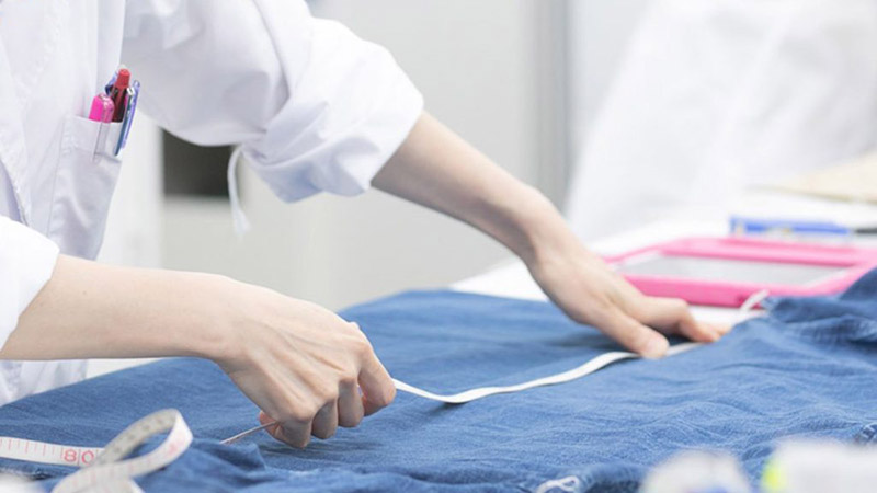 A person in a white lab coat is measuring a piece of blue fabric with a measuring tape on a worktable.