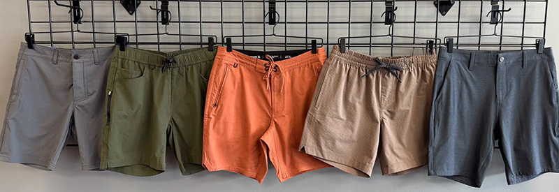 Five pairs of men's shorts hanging on a black grid wall. From left to right: light gray, olive green, burnt orange, tan, and dark gray. The shorts feature a mix of buttoned and elastic waistbands, some with drawstrings, designed for casual and active wear.
