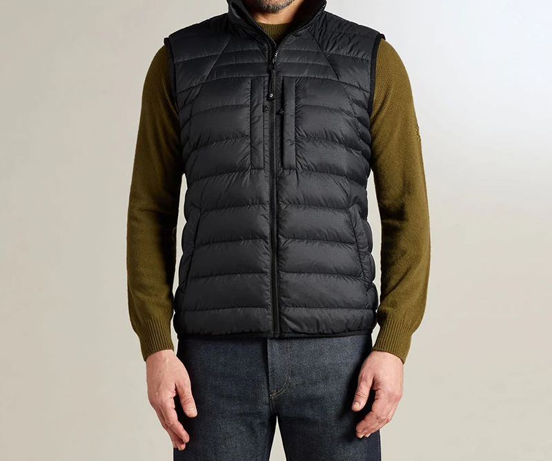 Man wearing a black quilted puffer vest over an olive long-sleeve sweater with dark jeans.