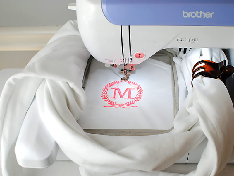 Embroidery machine stitching pink monogram design onto white fabric.