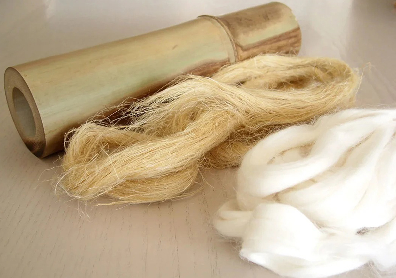 A bamboo segment lying on a surface next to two bundles of fibers: one light beige and coarse, the other white and soft, illustrating natural textile materials.