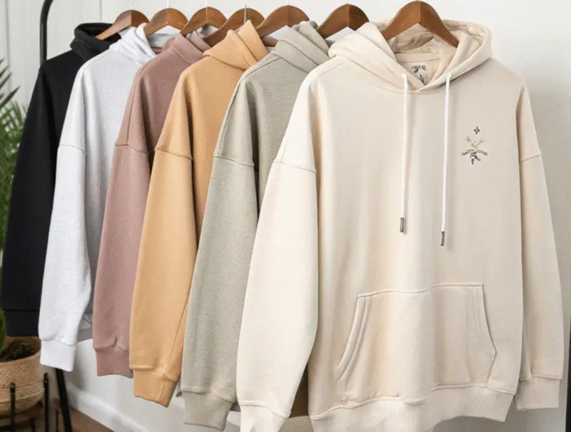 Oversized hoodies hanging in neutral tones including black, white, pink, beige, gray, and cream. Title: Neutral Hoodie Collection