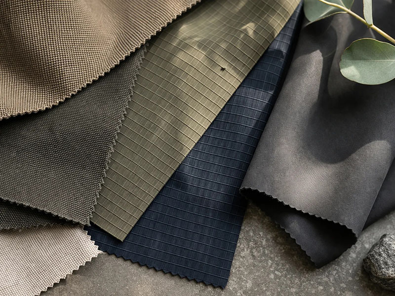 Various textured fabric swatches in earthy tones including brown, olive, navy, and black, laid out on a surface with a eucalyptus branch.