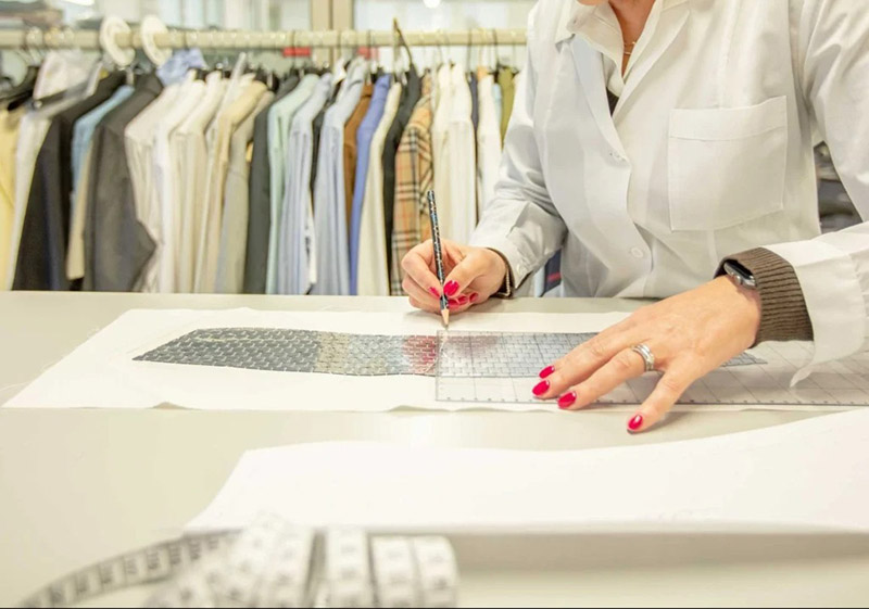 A person wearing a white lab coat is marking measurements on a clothing pattern using a ruler and pencil.