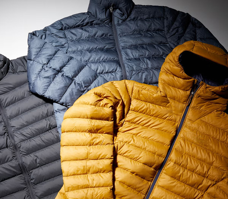 Three puffer jackets laid flat, overlapping each other, in charcoal gray, navy blue, and mustard yellow, highlighting their quilted design and full-length front zippers.
