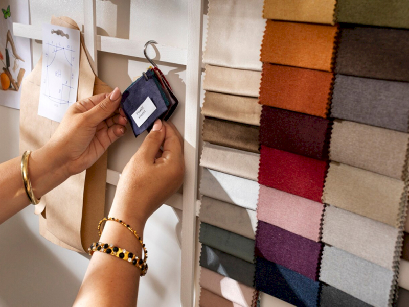 Hands holding a small fabric swatch book labeled “Textile Samples” in front of a pinned clothing pattern, next to a wall display of various rectangular fabric swatches in shades of beige, brown, red, orange, purple, and gray.