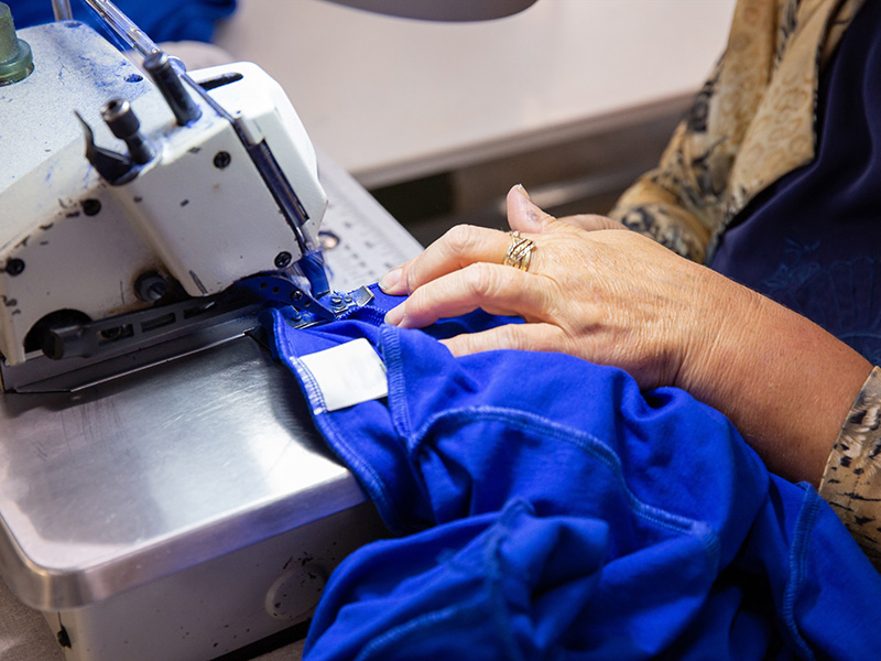 Close-up of a person's hands guiding a bright blue garment through an industrial sewing machine. The person is wearing rings, and the garment has a white label being stitched in place.