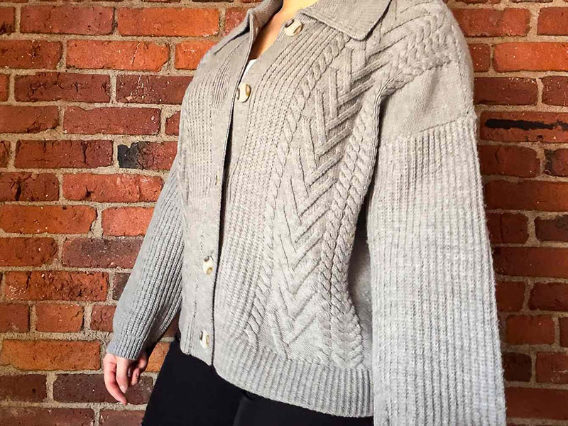 Woman wearing gray cable knit cardigan sweater standing against exposed brick wall