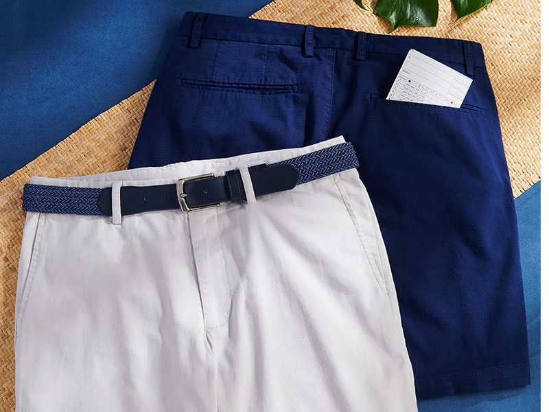 Pair of men's shorts, one white with blue belt in front, one navy blue with card in back pocket, laid on textured background.