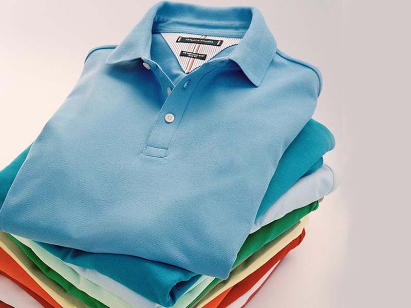 A neatly folded stack of polo shirts in various colors, with a light blue shirt on top featuring a small logo on the chest.