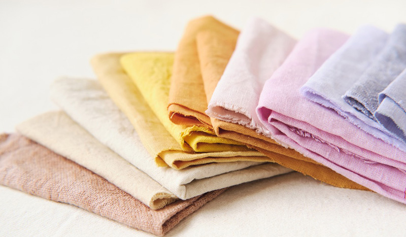 A neatly arranged stack of folded linen fabrics in soft pastel and earthy tones, including beige, yellow, orange, pink, and light blue, displayed on a light background.