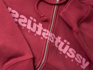 Close-up of red Stussy zip-up hoodie with bold logo print across chest and detailed stitching around zipper and cuffs