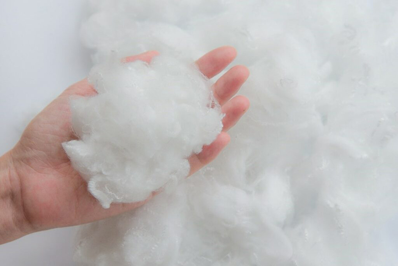 A hand holding a handful of white fluffy synthetic fiber, commonly used as stuffing for jackets, pillows, or cushions. More fiber is spread out in the background.