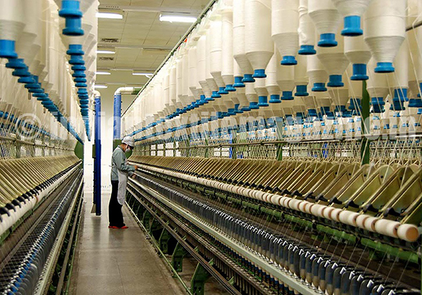 Worker monitoring a long row of spinning machines beneath white yarn cones in textile factory.