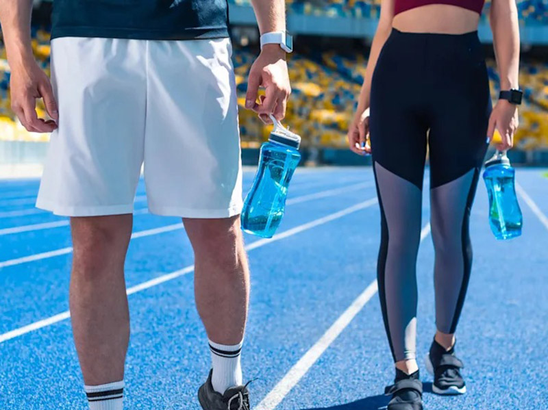 A man and a woman standing on a blue running track, each holding a clear blue water bottle.