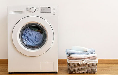 Front-loading washing machine beside a basket of folded clothes on a wooden floor.