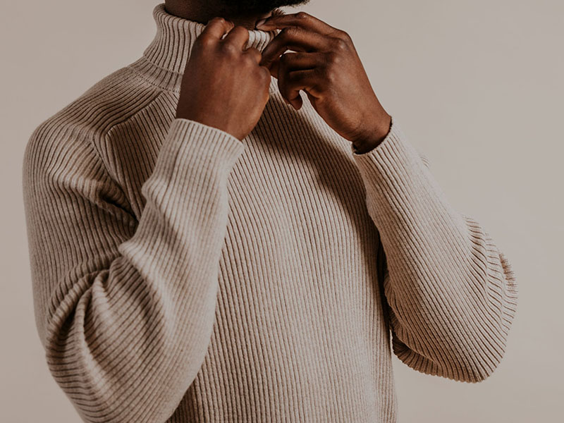 Person adjusting a beige ribbed turtleneck sweater, showing texture and long sleeves.