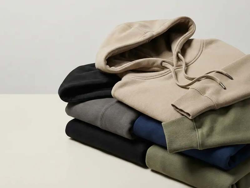 Stack of folded hooded sweatshirts in neutral colors including beige, black, gray, navy, and olive green on a plain surface.