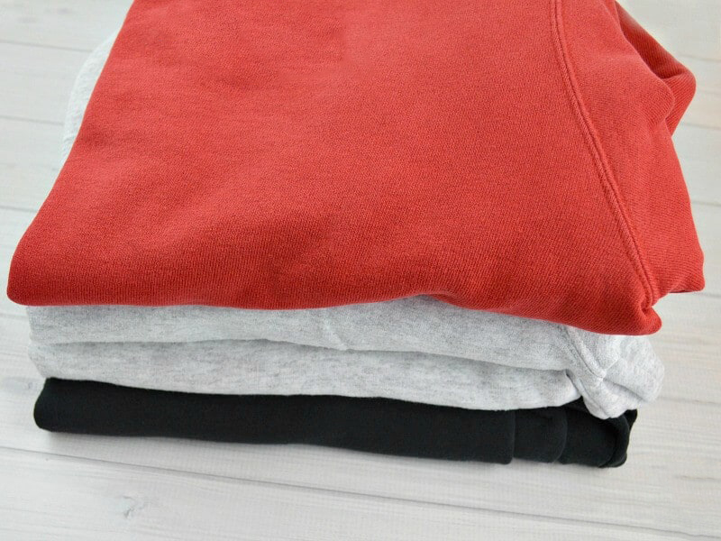 Stack of folded sweatshirts in coral, gray, and black casual fleece layers.