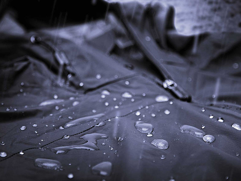 Close-up of a dark waterproof jacket with water droplets beading on the surface, showing water resistance in rain.