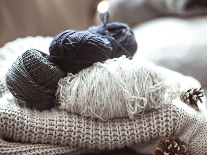 Three skeins of yarn in black, dark gray, and white resting on a beige knitted blanket with pinecones nearby.