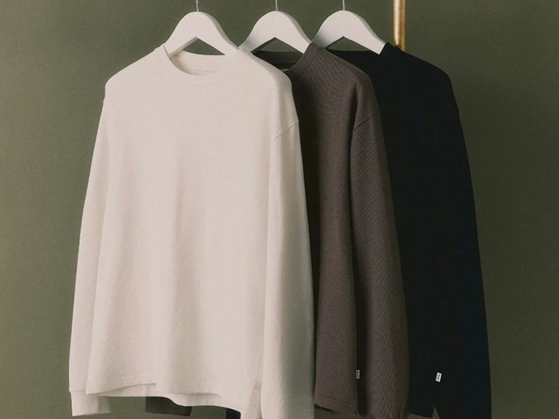 Three minimalistic sweatshirts hanging on wooden hangers, in white, brown, and black colors.