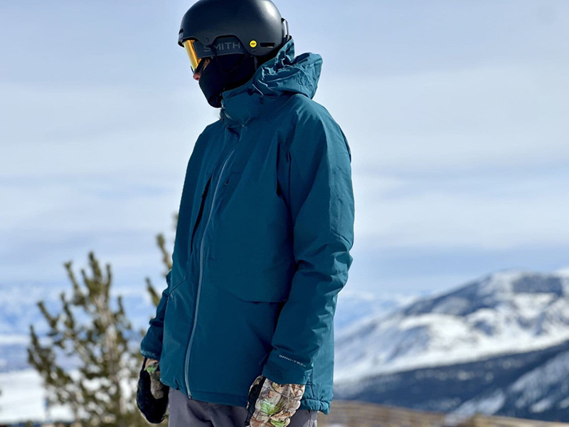 Person wearing blue insulated ski jacket with helmet and goggles in snowy mountain setting.