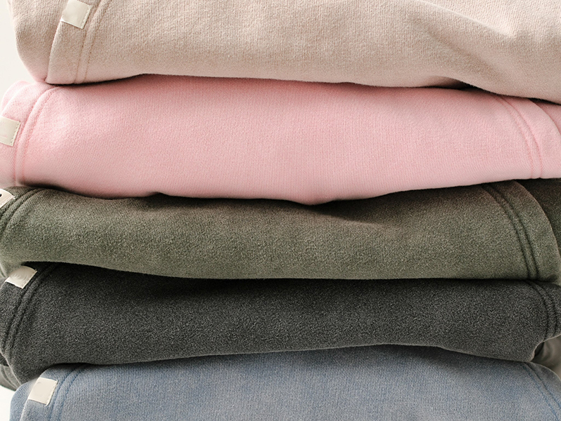 A neatly stacked pile of folded sweatshirts in five different colors: beige, pink, olive green, dark gray, and light blue. Each sweatshirt has a small white tag visible near the hem.