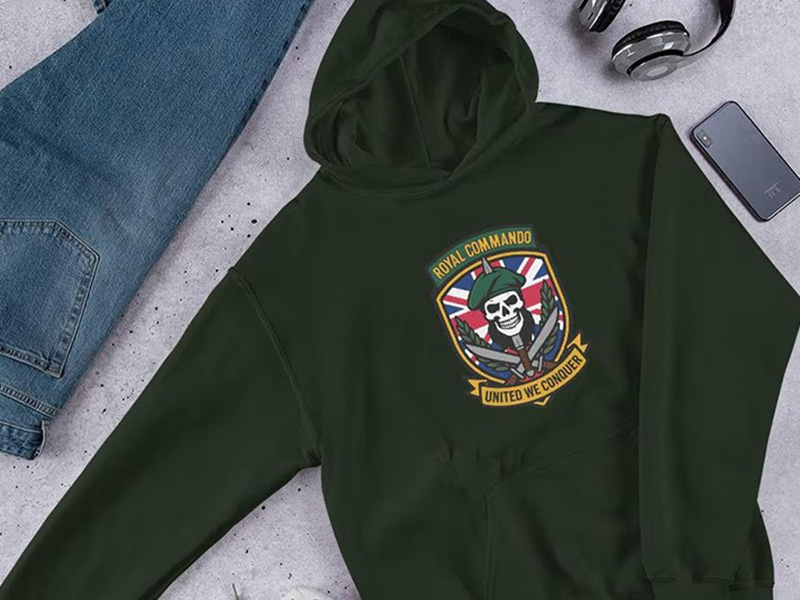 Dark green hoodie with military-style graphic print laid flat beside jeans and accessories.