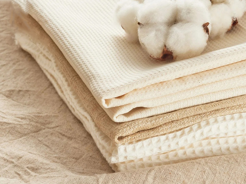 Folded cream and beige waffle-knit fabrics stacked with cotton bolls on top.