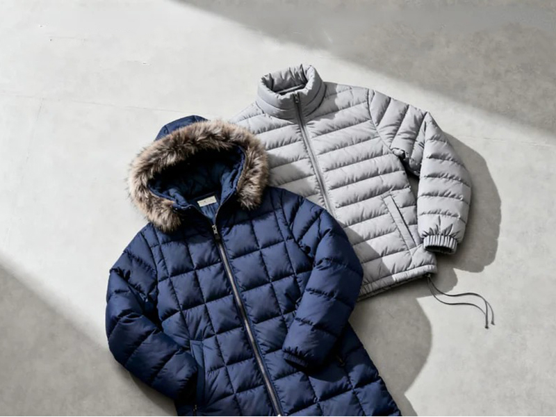 Two puffer jackets laid flat on a concrete surface, one navy blue with a fur-lined hood and the other light gray with a high collar, showcasing their quilted design and insulation.