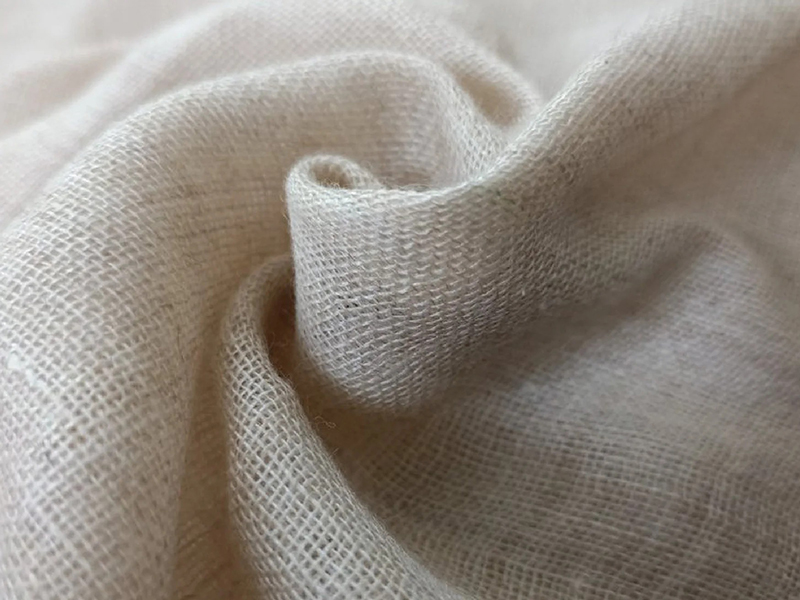 A detailed close-up image of a soft, beige-colored woven fabric, showing its fine texture and gentle folds.
