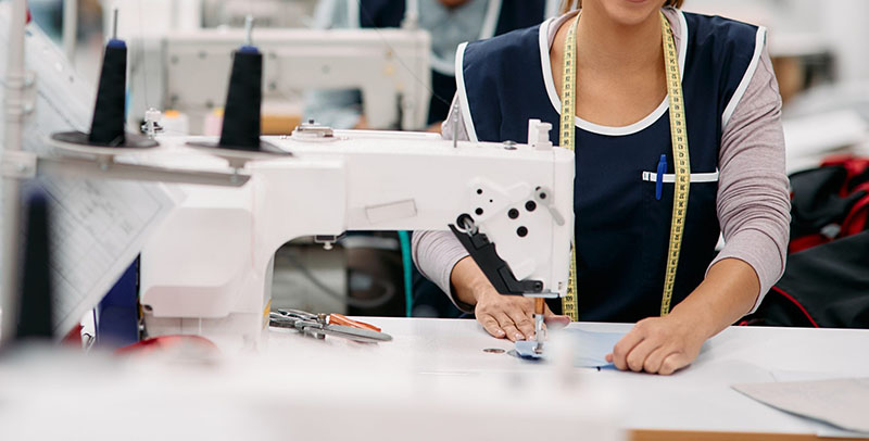 Close-up of a person using an industrial sewing machine, guiding fabric with hands. A measuring tape is draped around their neck in a bright, organized garment workshop.